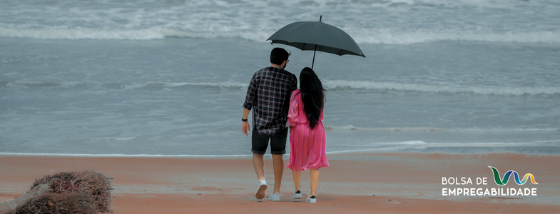 Imagem de um casal a passear na praia com um guarda-chuva, porque o tempo está frio e chuvoso. A imagem representa que é possível combater a sazonalidade no turismo e atrair turistas mesmo durante a época baixa através de estratégias práticas que vamos abordar de seguida.